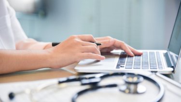 A person's hands typing on a laptop keyboard while a stethoscope is placed on top of the table