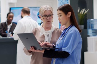 Senior woman and medical assistant analyzing diagnosis report on laptop, doing consultation in hospital reception. Nurse and patient discussing about treatment and health support. Handheld shot.