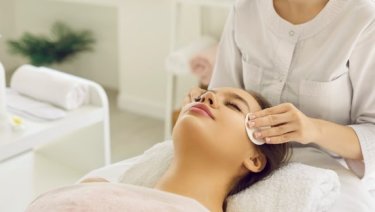 Woman enjoying a spa facial treatment, lying on table with eyes closed, towel on hair, surrounded by beauty products